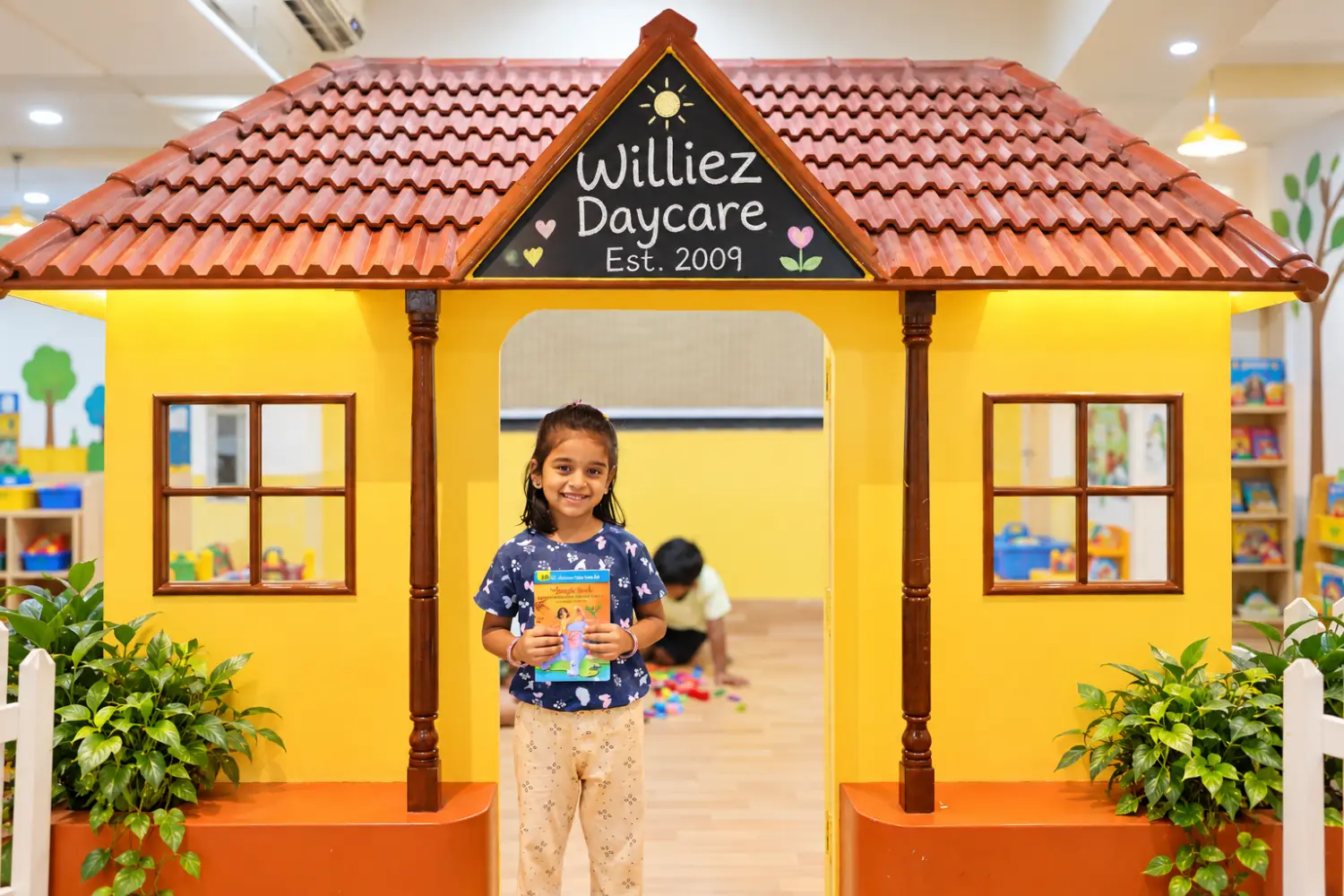 Smiling girl standing at the doorway of the Williez Daycare playhouse with potted plants